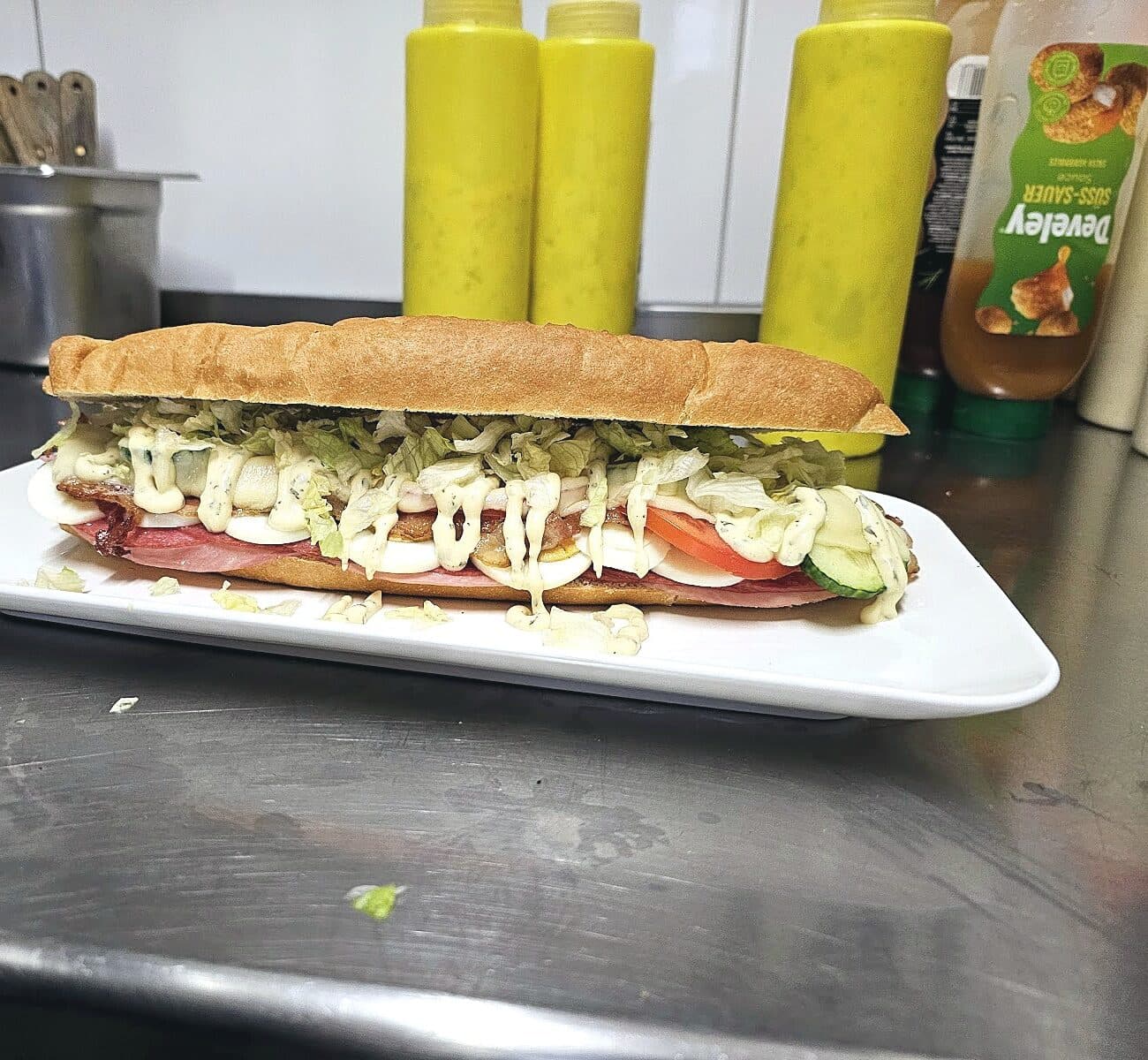 Langes Baguette, belegt mit Salat, Ei, Schinken, Tomate und heller Soße, auf einem weißen Teller