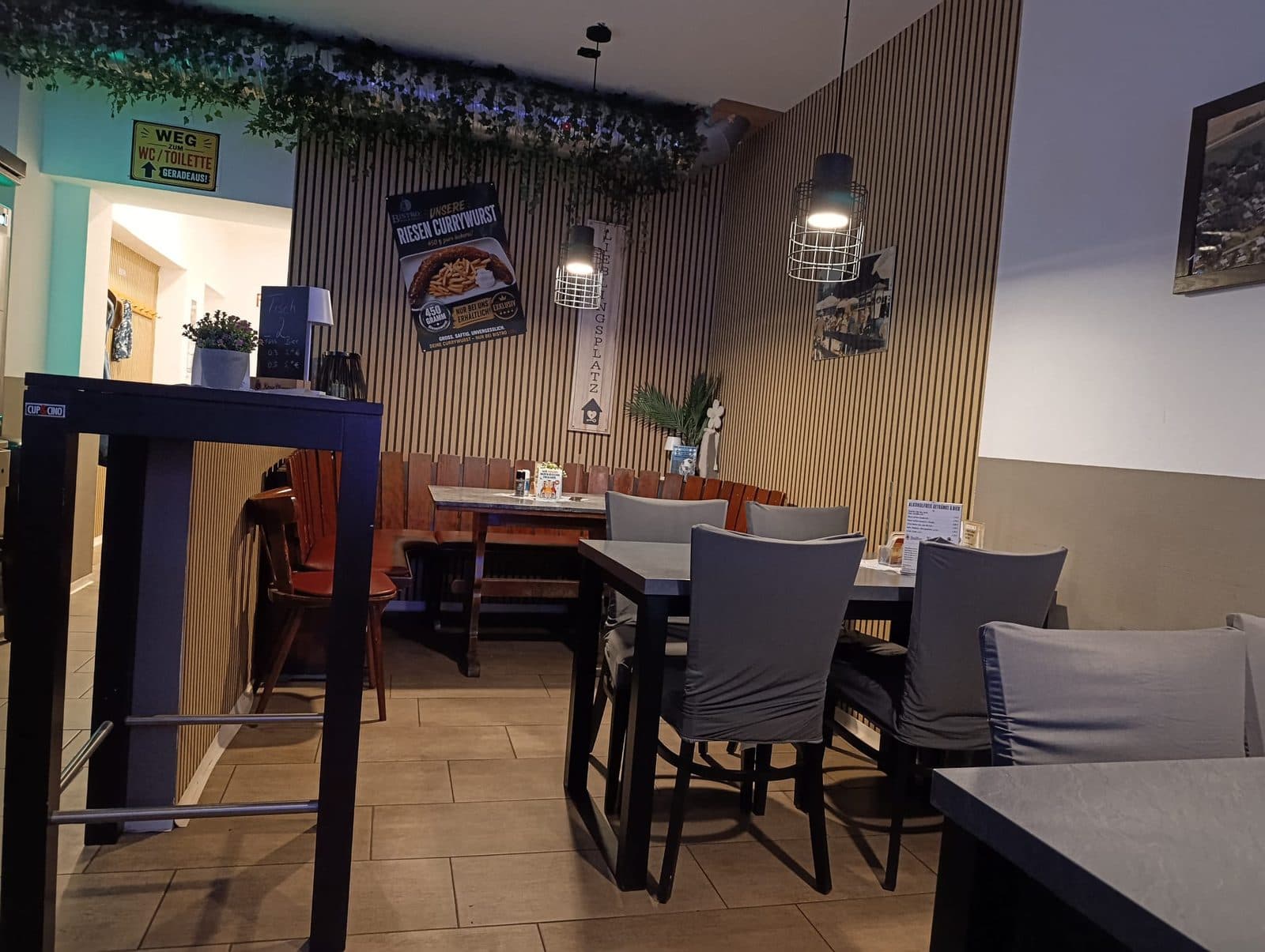 Gastraum mit Holzlattenwand, Efeu unter der Decke, Hochtisch und Tischen, an der Wand das Plakat «Riesen Currywurst»